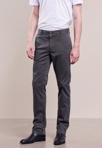 Man i mörkgrå slim-fit byxor, vit t-shirt instoppad, och svarta läderskor, stående mot en enkel beige bakgrund.