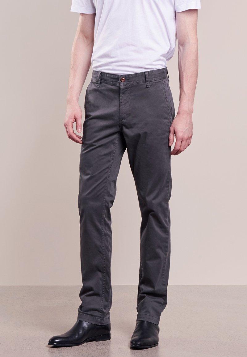 Man i mörkgrå slim-fit byxor, vit t-shirt instoppad, och svarta läderskor, stående mot en enkel beige bakgrund.