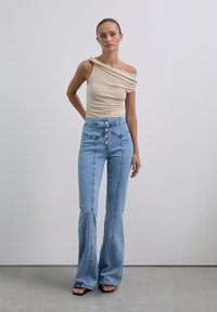 Vrouw die binnen staat in een beige top met één schouder, hooggewicht blauwe flared jeans en zwarte open teen sandalen tegen een vlakke muur.