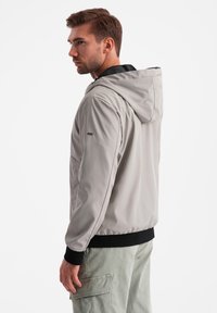 Giacca con cappuccio grigio chiaro dal texture liscia, con polsini elasticizzati e dettagli neri. Presenta una vestibilità rilassata e tasche angolate sui lati.