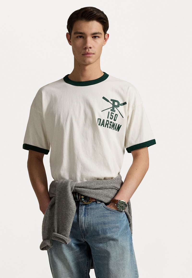 T-shirt blanc en coton avec col et poignets côtelés verts, arborant le texte "150 OARSMAN" et un graphique de rame croisées. Accompagné d'un pull gris noué à la taille.