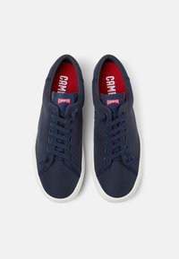 Zapatillas de lona azul marino con suelas de goma blanca, que presentan seis ojales, detalle en la puntera y un forro de tela roja con el logotipo.