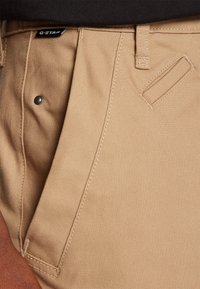 Pantalones de algodón beige con un botón visible, bolsillos frontales en ángulo y etiqueta de marca. La textura es suave con un diseño ligeramente estructurado.