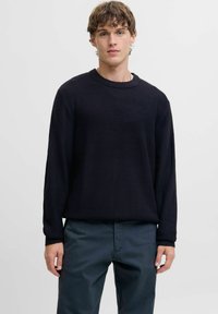 Suéter de punto acanalado en azul marino oscuro con mangas largas y cuello redondo; combinado con pantalones oscuros de ajuste relajado y patrón texturizado.