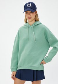 Sudadera verde menta con capucha ajustable, corte relajado y puños acanalados, combinada con una gorra azul marino que lleva un logo blanco de Harvard.