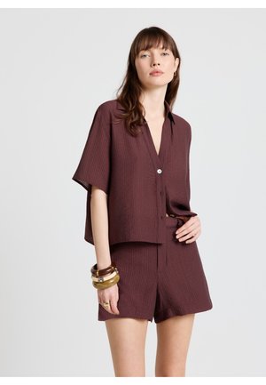 Femme portant une chemise bordeaux à manches courtes ample avec des boutons et un short assorti, accessoirisée de bracelets et bagues épaisses, sur un fond uni.