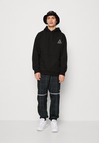 Sort hætte sweatshirt med frontlomme og trekantet logo, parret med grønternet bukser med hvide striber. Hvide sneakers fuldender outfittet.