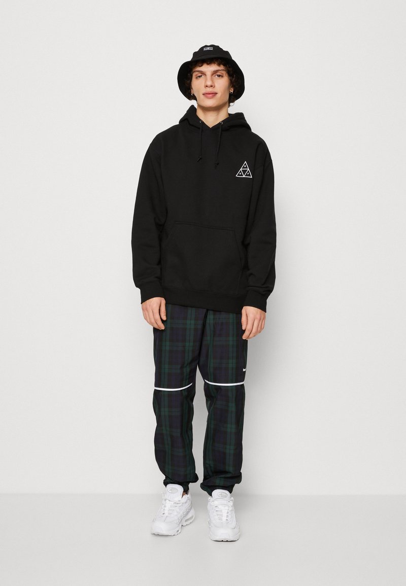 Sort hætte sweatshirt med frontlomme og trekantet logo, parret med grønternet bukser med hvide striber. Hvide sneakers fuldender outfittet.