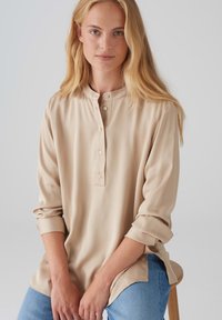 Beige Hemdbluse mit Stehkragen, langen Ärmeln und lockerer Passform, aus glattem Stoff mit einem dezenten Glanz.