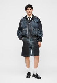 Jeune mannequin masculin portant une veste en jean noire et bleue avec quatre grandes poches avant, un short en jean assorti jusqu'aux genoux, des chaussettes blanches et des mocassins noirs.