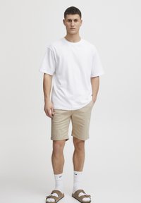 Weißes Baumwoll-T-Shirt, lockere Passform, beigefarbene Shorts, weiße Socken und braune Sandalen. Minimalistischer Stil ohne sichtbare Muster oder Logos.