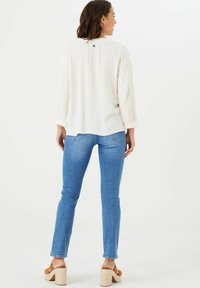 Blusa larga color beige claro de manga larga con escote redondo y un sutil detalle de botones en la parte trasera. Combinada con jeans ajustados azules y sandalias de plataforma color tan.