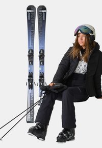 Skiequipment met blauwe en zwarte ski's met bindingen, en een persoon gekleed in een zwarte geïsoleerde jas, zwarte broeken en ski-boots.