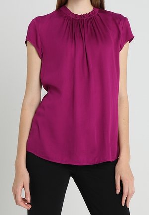 Kobieta w bezrękawnikowym, magentowym bluzce z falbanką na szyi i czarnych spodniach, stojąca na tle jednolitego jasnego tła.