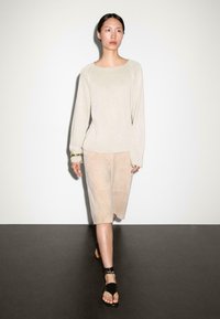 Maglione a maniche lunghe color beige chiaro con scollo rotondo, abbinato a pantaloni corti in suede beige. Il modello indossa sandali neri con fascetta alla caviglia.
