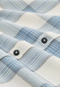 Blauw-witte geruite stof met een gestructureerde weving. Heeft zwarte knoopdetails. De patronen omvatten strepen en vierkanten in verschillende tinten.