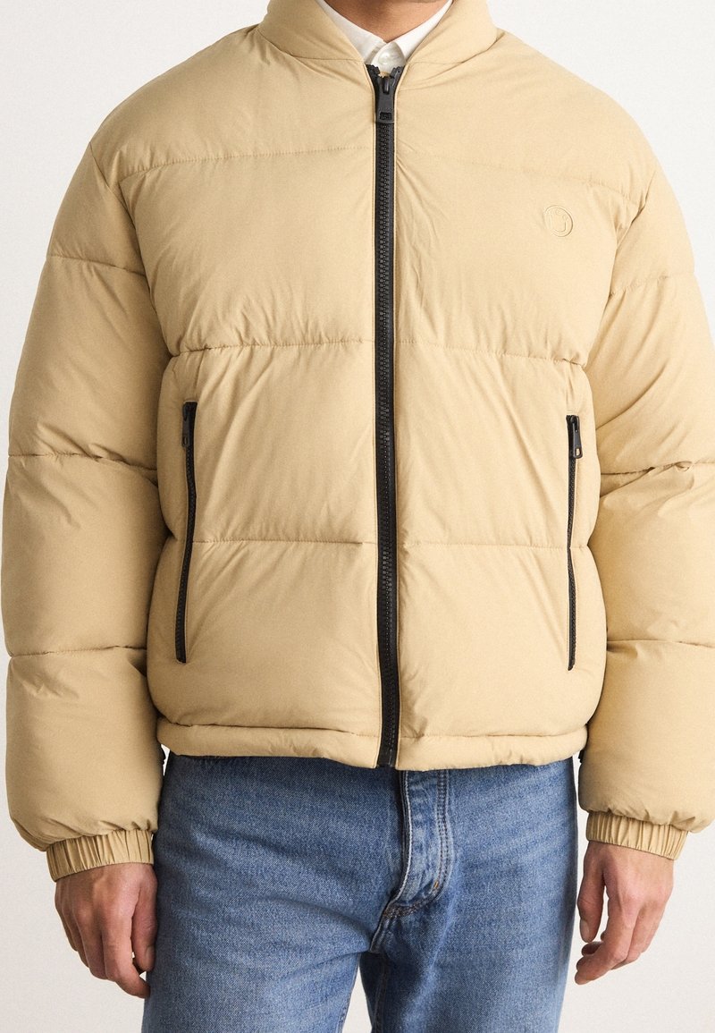 Vaalean beige puffer-takki, jossa on korkea kaulus, musta vetoketju ja vetoketjulliset sivutaskut. Ominaisuuksiin kuuluu kuvioitu pinta ja joustavat hihansuut.