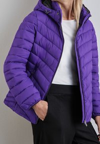 Kobieta w fioletowej pikowanej kurtce z kapturem, nosząca białą koszulę i czarne spodnie, z jedną ręką w kieszeni, stojąca przy szarej ścianie.