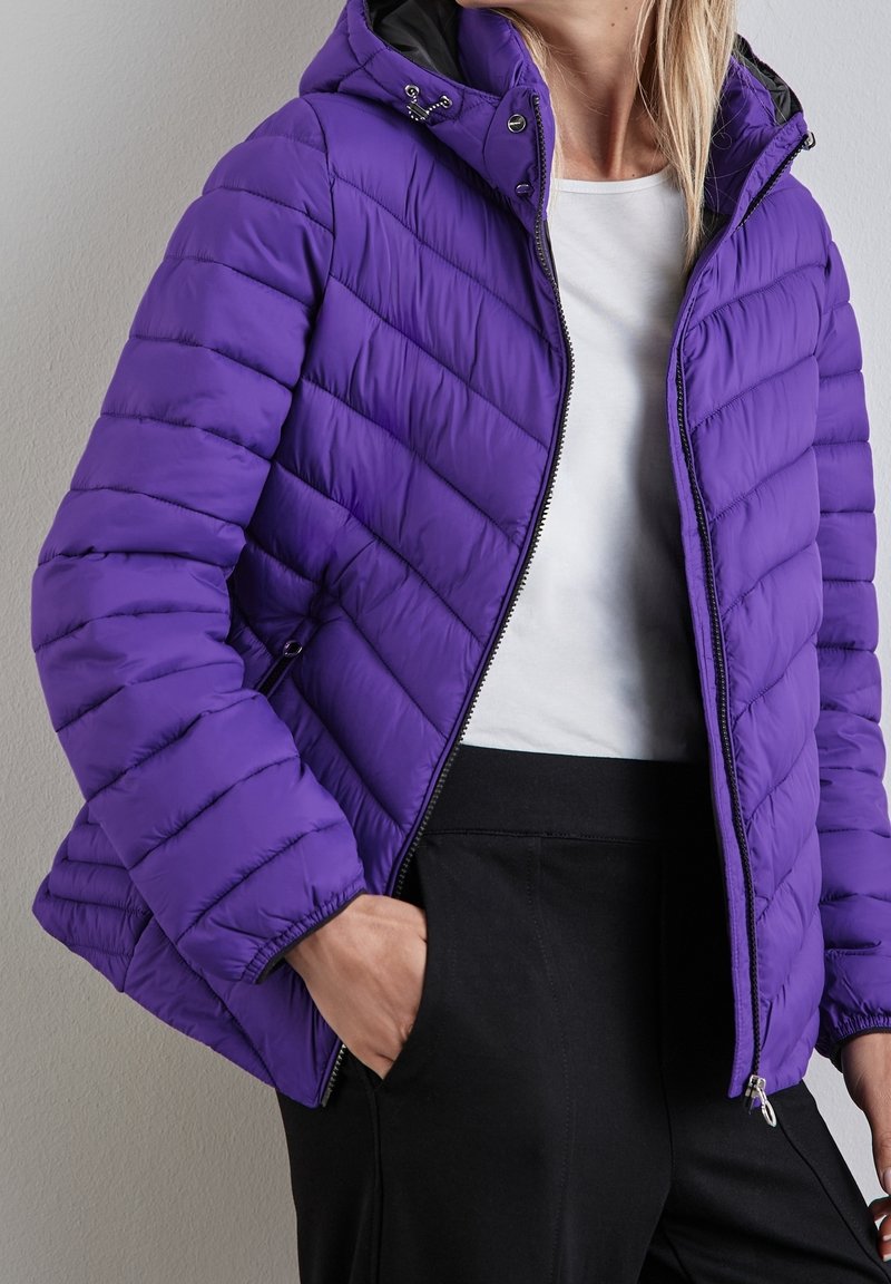 Kobieta w fioletowej pikowanej kurtce z kapturem, nosząca białą koszulę i czarne spodnie, z jedną ręką w kieszeni, stojąca przy szarej ścianie.