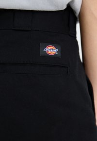 Pantaloni negri dintr-un material texturat, cu un buzunar la spate și o etichetă colorată cu logo-ul "Dickies". Design simplu și funcțional.
