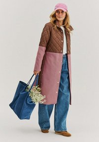 Hnědý prošívaný dlouhý kabát s růžovým houndstooth spodkem, spárovaný s kalhotami širokého střihu, růžovou čepicí a modrou taškou přepravující květiny.