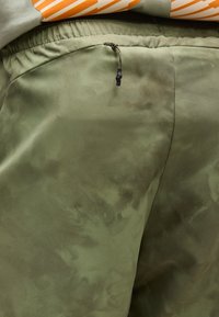 Κοντινό πλάνο ελιάς πράσινου παντελονιού με κορδόνι στη μέση και διακριτικά σκοτεινότερα σημεία, φορεμένο με λευκό πουκάμισο με πορτοκαλί ρίγες.