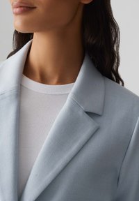 Blazer azzurro chiaro con rever a taglio, tessuto testurizzato, indossato sopra un top bianco a coste. Focus sui dettagli del colletto e del scollo.