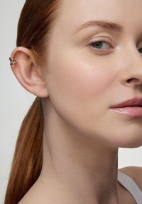Zbliżenie na osobę noszącą srebrny klipe, charakteryzujący się geometrycznym wzornictwem. Kształt cery jest jasny, a włosy są proste i czerwone.