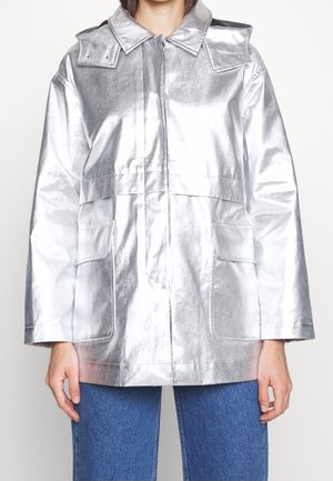 Osoba ubrana w błyszczącą srebrną metaliczną kurtkę z dużymi przednimi kieszeniami i niebieskie dżinsy, stojąca na tle jednolitego białego tła.