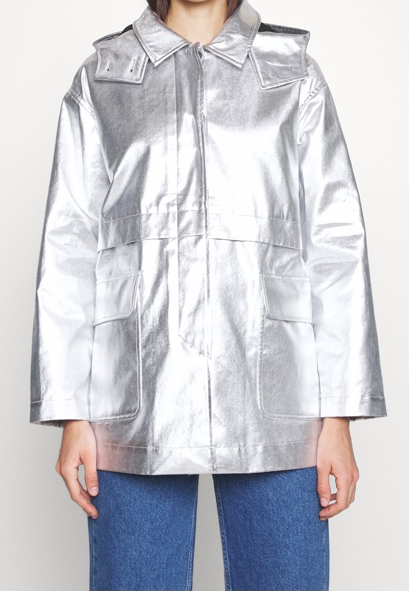 Personne portant une veste métallique argentée brillante avec de grandes poches avant et un jean bleu, debout devant un fond blanc uni.