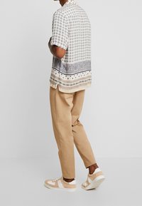 Valkoinen napillinen paita, jossa on ruudullinen kuvio ja koristeellisia sinisiä yksityiskohtia. Yhdistetty vaaleanruskeisiin housuihin ja kermaisiin tennareihin.