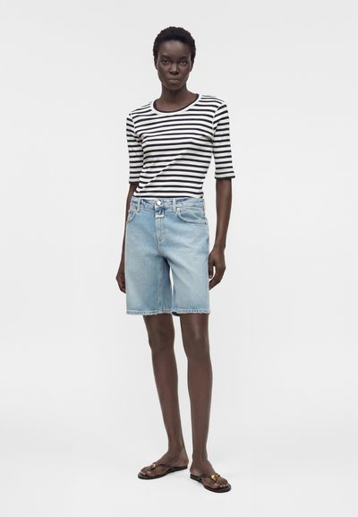 Γυναίκα που στέκεται φορώντας μια μαύρο-λευκή ριγέ μπλούζα με μανίκια μέχρι τη μέση του βραχίονα, ανοιχτό μπλε τζιν σορτς και μαύρα σανδάλια με χρυσές λεπτομέρειες.