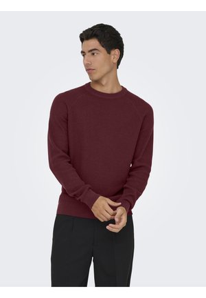 Suéter de punto burdeos con cuello redondo, de mangas largas y con bajo acanalado. El modelo lleva pantalones oscuros, con las manos colocadas en la cintura.