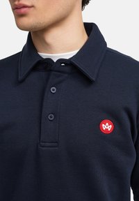 Polo shirt blu navy con colletto, tre bottoni e un logo ricamato rosso sul petto. Materiale in cotone con una texture liscia.