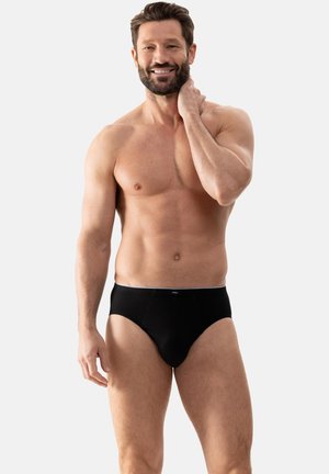 Uomo senza maglietta con barba e capelli corti indossa slip neri, in piedi contro uno sfondo chiaro e semplice, sorridente e che si tocca il collo.