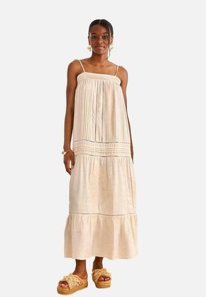 Mujer con un vestido largo color beige claro sin mangas, con pliegues y detalles de encaje, combinado con sandalias beige de plataforma con detalles de flecos.