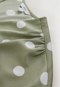 Tissu vert à pois blancs avec couture froncée et une étiquette de vêtement visible portant la mention "NEXT" sur un fond clair.