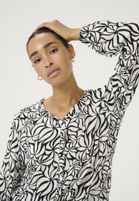Black and white patterned blouse with a ruffled neckline, long sleeves, and a relaxed fit; texture appears lightweight and crinkled.