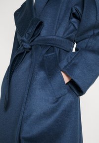 Personne portant un manteau en laine bleu foncé avec une ceinture nouée, main dans la poche latérale, montrant la texture et les détails du design du manteau.