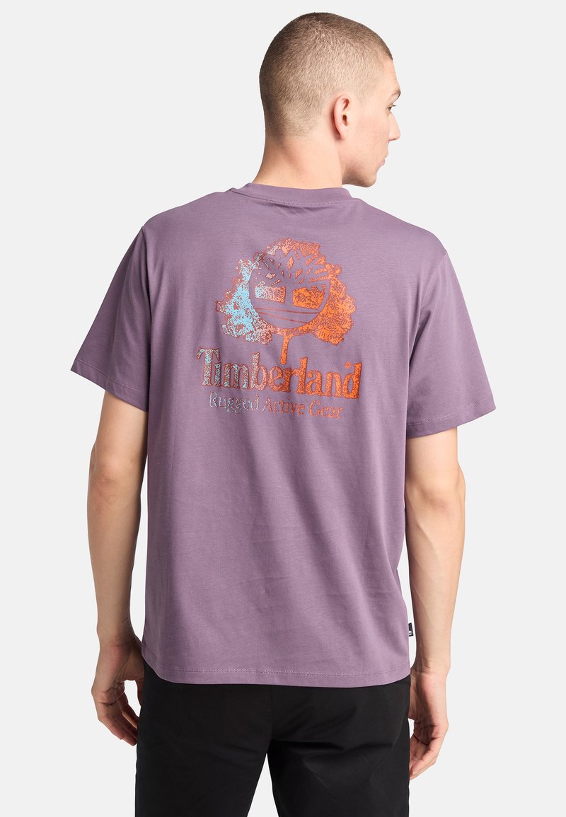 Camiseta de algodón morada con un gran logo de árbol y el texto "Timberland" en metálico naranja y azul, con un corte casual y mangas cortas.