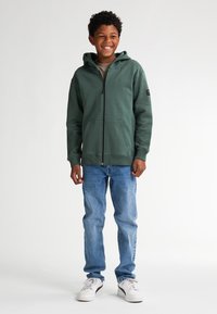 Groene ritshoodie met een voorzak en verstelbare capuchon, in combinatie met lichtblauwe jeans en witte sneakers. Eenvoudig ontwerp en zachte textuur.