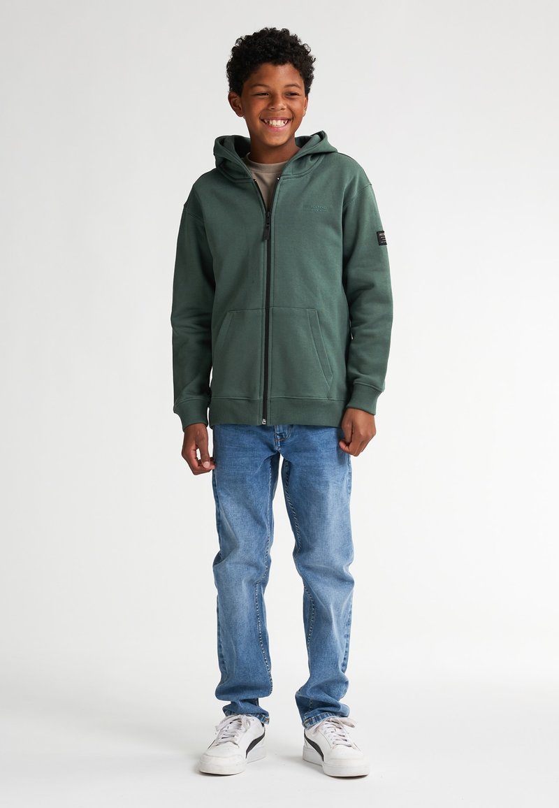 Groene ritshoodie met een voorzak en verstelbare capuchon, in combinatie met lichtblauwe jeans en witte sneakers. Eenvoudig ontwerp en zachte textuur.