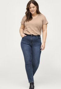 Maglietta a maniche corte marrone chiaro abbinata a jeans blu scuro a vita alta. I jeans presentano un classico design a cinque tasche e una leggera elasticità.
