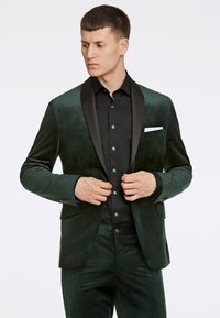 Blazer in velluto verde scuro con rever in satin, abbinato a una camicia nera con bottoni. Texture notevoli e dettagli di design eleganti.