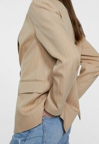 Beige kavaj med smala ränder, en ficka, skräddarsydd passform och strukturerade axlar, matchad med ljusblå slitna jeans.