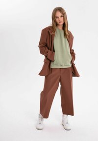 Brun oversized blazer över en ljusgrön sweatshirt, kombinerad med bruna vida byxor och vita sneakers. Tyget verkar vara glatt.