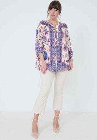 Witte bloeiende blouse met kleurrijke patronen en een blauwe bies, gecombineerd met lichtbeige cropped broeken en nude hakken. Lichtgewicht stof.