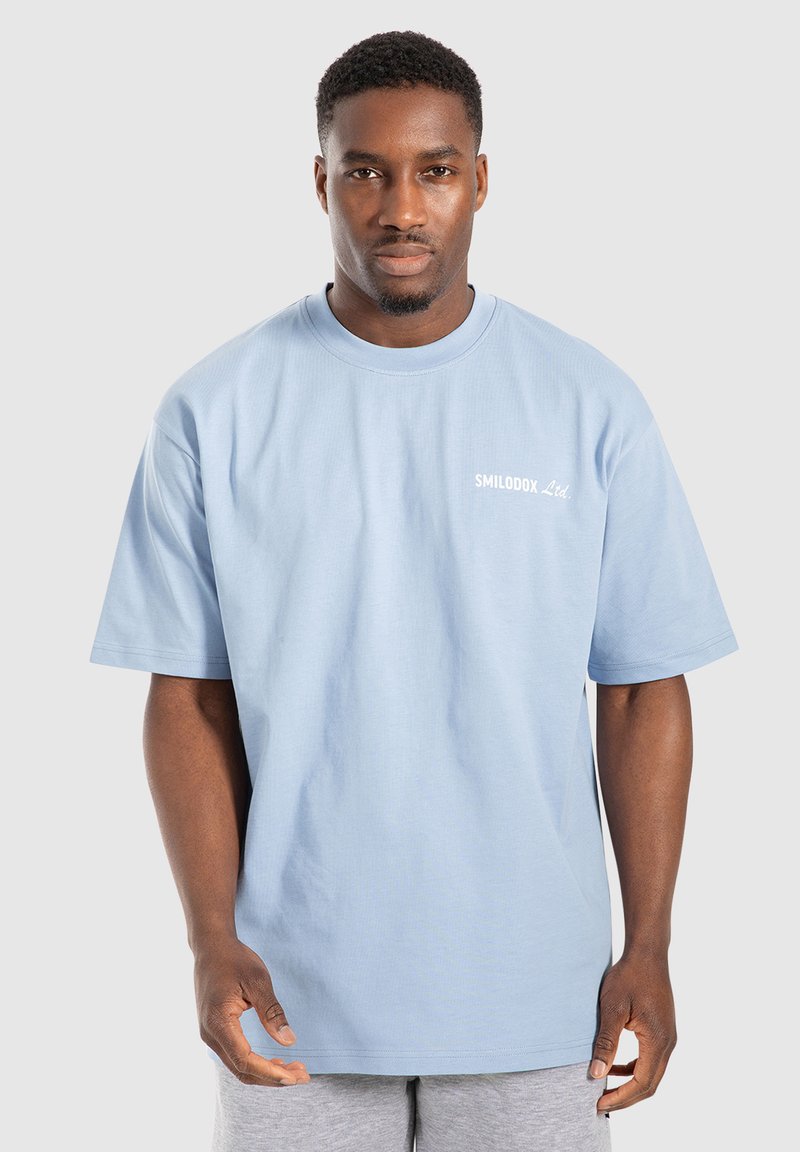 Camiseta de algodón azul claro con un corte holgado, cuello redondo acanalado, mangas cortas y el logo blanco "SMILODOX Ltd." en el lado izquierdo del pecho.