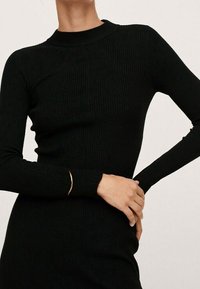 Femme portant une robe noire ajustée à manches longues en côtes, posant avec un bras fléchi et l'autre main reposant sur sa hanche.