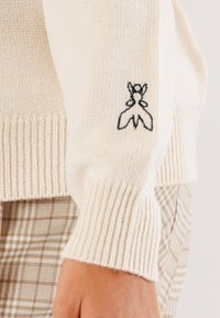Maglione in crema a maglia con polsini a costine, caratterizzato da un design ricamato nero di una farfalla sulla manica. La texture è morbida e liscia.